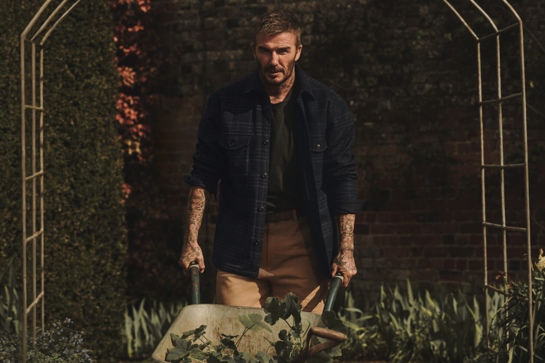 David Beckham in een donkerblauw geruit overshirt, zwart T-shirt en kakikleurige broek in een Engelse tuin