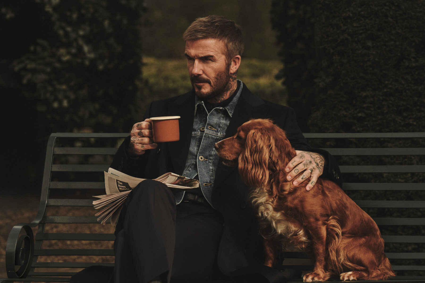 David Beckham op een bankje met een mok koffie en een hond, in een zwarte blazer over een spijkerjasje