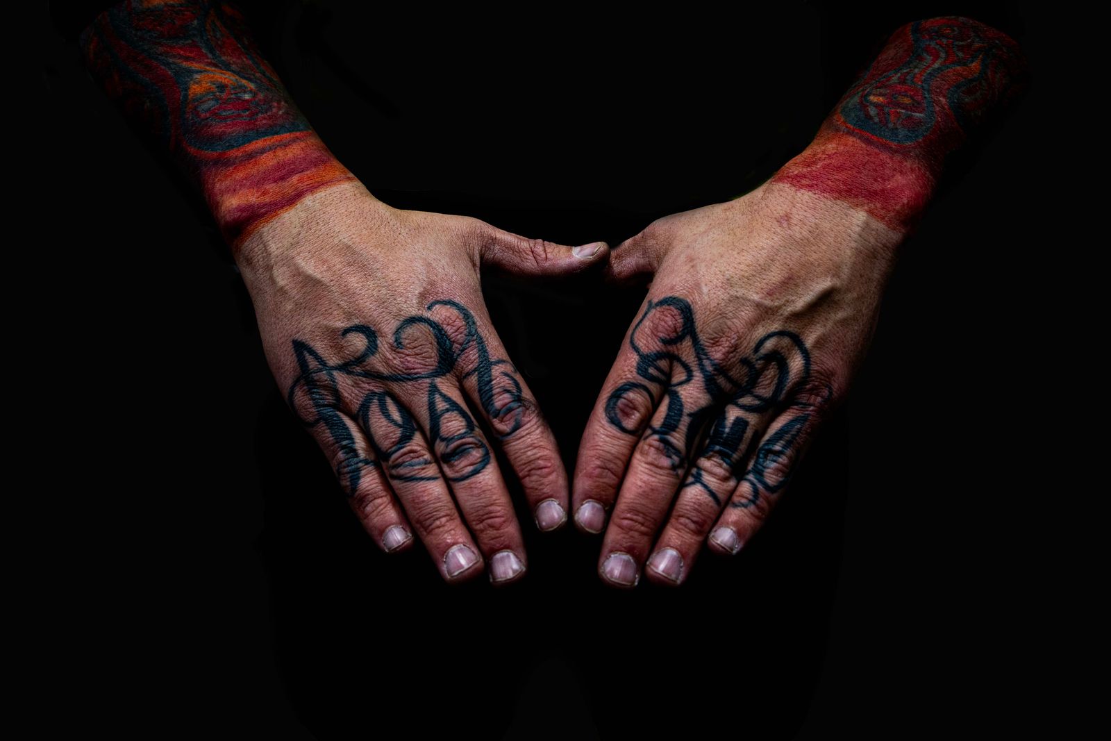 Twee handen met getatoeëerde letters op de vingers en kleurrijke tattoos op de polsen tegen een zwarte achtergrond