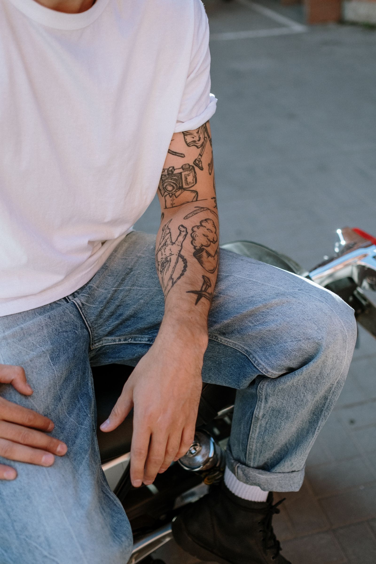 Man in een wit T-shirt en spijkerbroek met meerdere zwarte tattoos op zijn onderarm, zittend op een motor