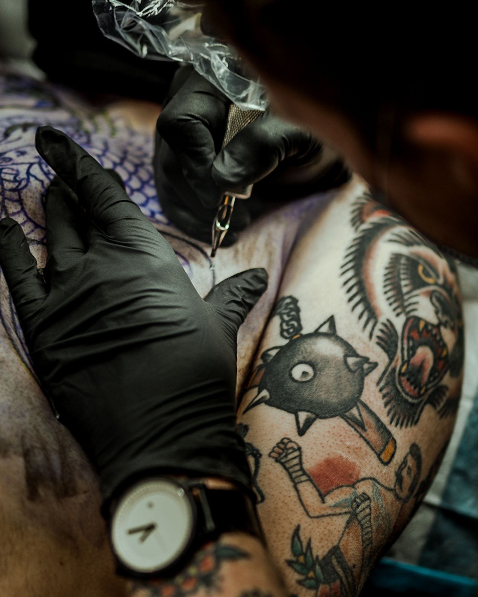 Tatoeëerder met zwarte handschoenen werkt aan een traditionele tattoo met dikke zwarte lijnen en rode accenten op een arm